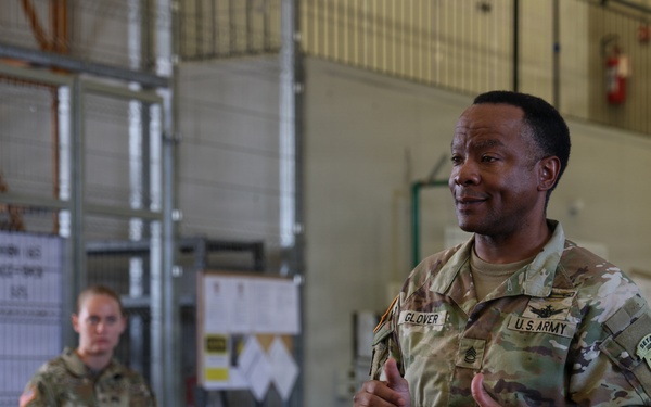 A U.S. Army soldier named Glover, identifiable by name tape and rank insignia, appears to be addressing a small group in what looks like a military hangar or warehouse facility. He is gesturing with both hands in a thumbs-up position, suggesting an instructional or motivational briefing. At least two other uniformed soldiers are visible — one seated and one standing in the background.