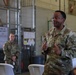 A U.S. Army soldier named Glover, identifiable by name tape and rank insignia, appears to be addressing a small group in what looks like a military hangar or warehouse facility. He is gesturing with both hands in a thumbs-up position, suggesting an instructional or motivational briefing. At least two other uniformed soldiers are visible — one seated and one standing in the background.