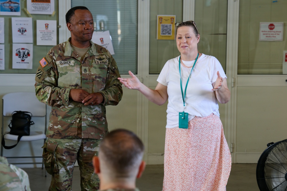 A U.S. Army soldier named Glover, wearing OCP camouflage uniform, stands alongside a civilian woman in casual attire who appears to be speaking or presenting to a small audience. The setting appears to be a military facility or medical area, indicated by posted signs and notices on the walls behind them. Both individuals appear engaged in a briefing or informational session.