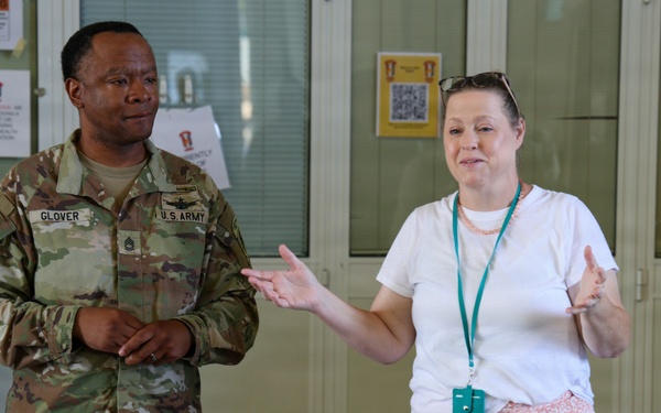 A U.S. Army soldier named Glover, wearing OCP camouflage uniform, stands alongside a civilian woman in casual attire who appears to be speaking or presenting to a small audience. The setting appears to be a military facility or medical area, indicated by posted signs and notices on the walls behind them. Both individuals appear engaged in a briefing or informational session.