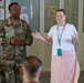 A U.S. Army soldier named Glover, wearing OCP camouflage uniform, stands alongside a civilian woman in casual attire who appears to be speaking or presenting to a small audience. The setting appears to be a military facility or medical area, indicated by posted signs and notices on the walls behind them. Both individuals appear engaged in a briefing or informational session.
