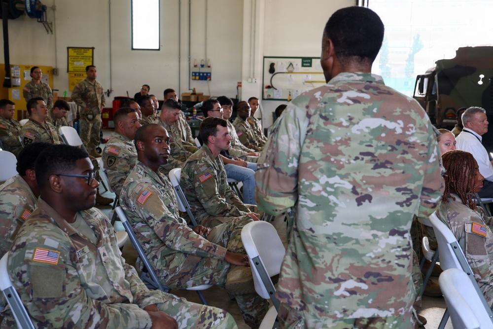 A group of U.S. Army soldiers in Operational Camouflage Pattern (OCP) uniforms are seated in folding chairs inside what appears to be a military maintenance bay or hangar facility. A standing soldier, seen from behind, appears to be addressing or briefing the assembled group. The setting suggests a formal unit briefing, training session, or command address.