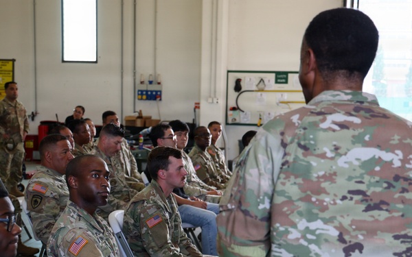 A group of U.S. Army soldiers in Operational Camouflage Pattern (OCP) uniforms are seated in folding chairs inside what appears to be a military maintenance bay or hangar facility. A standing soldier, seen from behind, appears to be addressing or briefing the assembled group. The setting suggests a formal unit briefing, training session, or command address.