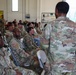 A group of U.S. Army soldiers in Operational Camouflage Pattern (OCP) uniforms are seated in folding chairs inside what appears to be a military maintenance bay or hangar facility. A standing soldier, seen from behind, appears to be addressing or briefing the assembled group. The setting suggests a formal unit briefing, training session, or command address.