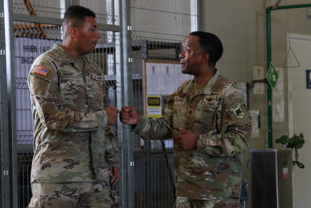 Two U.S. Army soldiers in OCP (Operational Camouflage Pattern) uniforms are engaged in a fist bump greeting inside what appears to be a military facility. The soldier on the left has a name tape reading 'GREBERRY' and the soldier on the right has a name tape reading 'GLOVER', with a SETAF-AF (U.S. Army Africa) patch visible on his right shoulder. The interaction appears friendly and informal.