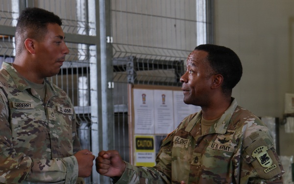 Two U.S. Army soldiers in OCP (Operational Camouflage Pattern) uniforms are engaged in a fist bump greeting inside what appears to be a military facility. The soldier on the left has a name tape reading 'GREBERRY' and the soldier on the right has a name tape reading 'GLOVER', with a SETAF-AF (U.S. Army Africa) patch visible on his right shoulder. The interaction appears friendly and informal.