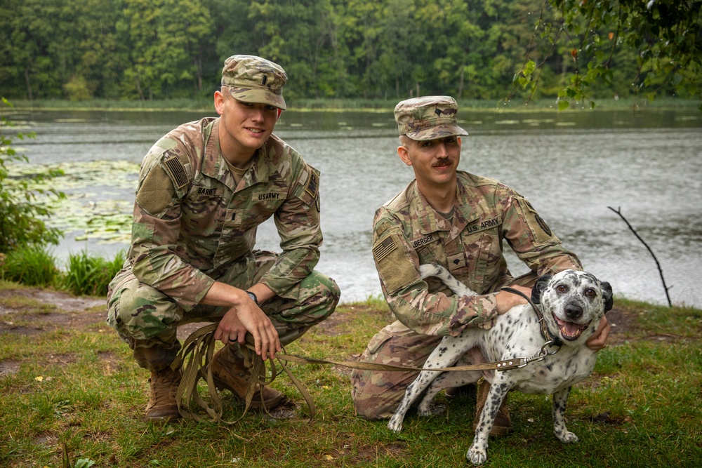 Two U.S. Army soldiers in OCP (Operational Camouflage Pattern) uniforms are crouching beside a lake with a Dalmatian dog on a leash. The soldiers are smiling and posing for the photo in what appears to be a relaxed, informal setting near a body of water surrounded by trees. Name tapes are partially visible on both uniforms, with one reading 'BARRY' and the other appearing to read 'BERGER'.