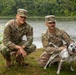 Two U.S. Army soldiers in OCP (Operational Camouflage Pattern) uniforms are crouching beside a lake with a Dalmatian dog on a leash. The soldiers are smiling and posing for the photo in what appears to be a relaxed, informal setting near a body of water surrounded by trees. Name tapes are partially visible on both uniforms, with one reading 'BARRY' and the other appearing to read 'BERGER'.