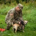 A U.S. Army soldier in MultiCam (OCP) camouflage uniform is crouched down on grass attending to a small tan/beige dog on a black leash. The soldier is wearing a patrol cap and has a U.S. flag patch visible on the left sleeve. A red object, possibly a bag or wrapper, is visible on the ground nearby.