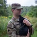 A U.S. Army soldier in OCP (Operational Camouflage Pattern) uniform is holding a small black cat or kitten in an outdoor, vegetated setting. The soldier is looking down at the animal with a calm expression. The background consists of lush green foliage and overcast skies.