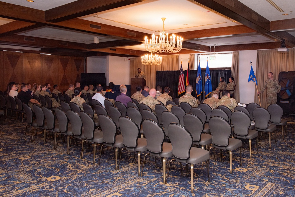 A military ceremony or briefing is taking place in a formal banquet or conference room with wood-paneled walls and a chandelier. Personnel in military camouflage uniforms (appearing to be U.S. Air Force OCP/ABU pattern) are seated in rows of gray chairs facing a front podium area. Several flags are displayed at the front, including what appears to be a U.S. flag, a German flag, and U.S. Air Force unit guidons.