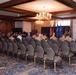 A military ceremony or briefing is taking place in a formal banquet or conference room with wood-paneled walls and a chandelier. Personnel in military camouflage uniforms (appearing to be U.S. Air Force OCP/ABU pattern) are seated in rows of gray chairs facing a front podium area. Several flags are displayed at the front, including what appears to be a U.S. flag, a German flag, and U.S. Air Force unit guidons.