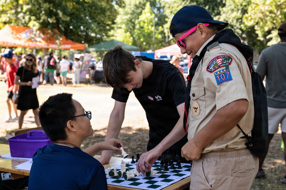 Three individuals are gathered around a chess board at an outdoor community event or fair. A Boy Scout wearing a Troop 107 uniform and pink sunglasses is smiling while engaging with a person in a black USA-branded shirt who is leaning over the chess board, and a seated person in a navy shirt observes. The setting appears to be a park with vendor tents and crowds visible in the background.
