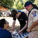 Three individuals are gathered around a chess board at an outdoor community event or fair. A Boy Scout wearing a Troop 107 uniform and pink sunglasses is smiling while engaging with a person in a black USA-branded shirt who is leaning over the chess board, and a seated person in a navy shirt observes. The setting appears to be a park with vendor tents and crowds visible in the background.