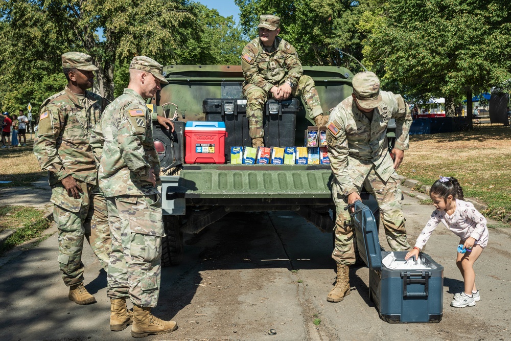 Four U.S. Army soldiers in OCP (Operational Camouflage Pattern) uniforms are unloading supplies from the back of a military HMMWV (Humvee). A young girl is assisting or interacting with one of the soldiers near a large dark cooler on the ground. The scene appears to be a community outreach or disaster relief distribution event in a park-like setting.