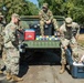 Four U.S. Army soldiers in OCP (Operational Camouflage Pattern) uniforms are unloading supplies from the back of a military HMMWV (Humvee). A young girl is assisting or interacting with one of the soldiers near a large dark cooler on the ground. The scene appears to be a community outreach or disaster relief distribution event in a park-like setting.