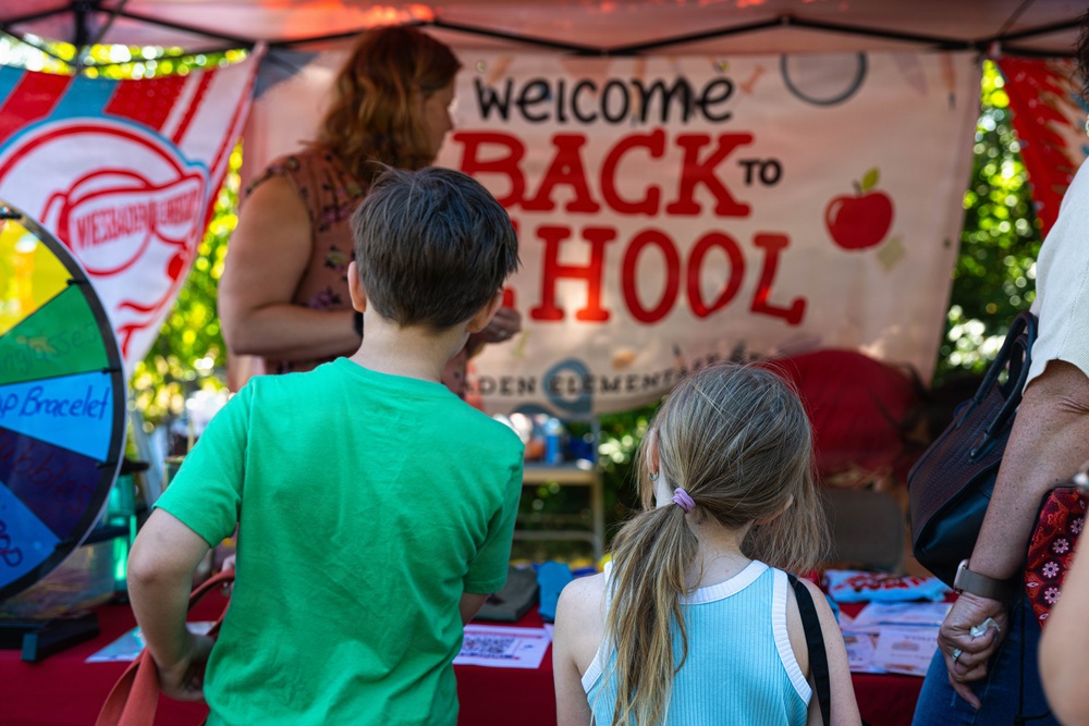 Two children are standing at an outdoor booth decorated with a 'Welcome Back to School' banner, likely at a community or school event. An adult woman stands behind the booth table, which is covered in red and features various items including a colorful prize wheel. The setting appears to be a back-to-school fair held under a canopy tent.