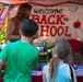 Two children are standing at an outdoor booth decorated with a 'Welcome Back to School' banner, likely at a community or school event. An adult woman stands behind the booth table, which is covered in red and features various items including a colorful prize wheel. The setting appears to be a back-to-school fair held under a canopy tent.