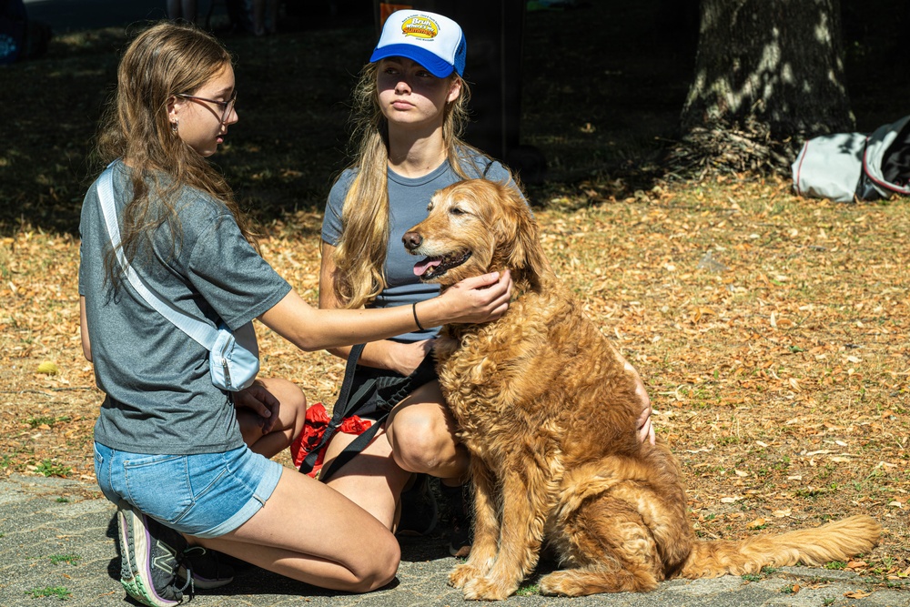 Two young teenage girls are interacting with a golden retriever dog outdoors in what appears to be a park or shaded outdoor area. One girl is kneeling and petting the dog while the other, wearing a blue and white trucker cap, is seated and holding the dog's leash. The dog appears calm and happy with its tongue out.