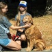Two young teenage girls are interacting with a golden retriever dog outdoors in what appears to be a park or shaded outdoor area. One girl is kneeling and petting the dog while the other, wearing a blue and white trucker cap, is seated and holding the dog's leash. The dog appears calm and happy with its tongue out.