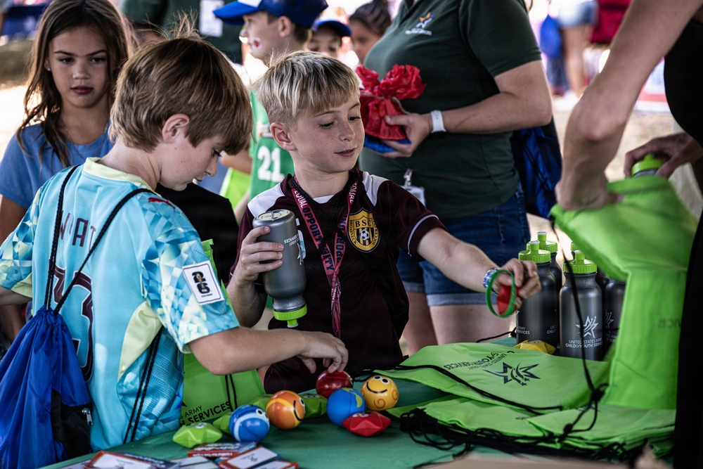 Two young boys are at a promotional booth for United States Army Child and Youth Services at what appears to be a youth sports event. They are picking up small colorful balls and examining items on a green-covered table filled with giveaway merchandise. Several adults and other children are visible in the background.