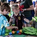 Two young boys are at a promotional booth for United States Army Child and Youth Services at what appears to be a youth sports event. They are picking up small colorful balls and examining items on a green-covered table filled with giveaway merchandise. Several adults and other children are visible in the background.