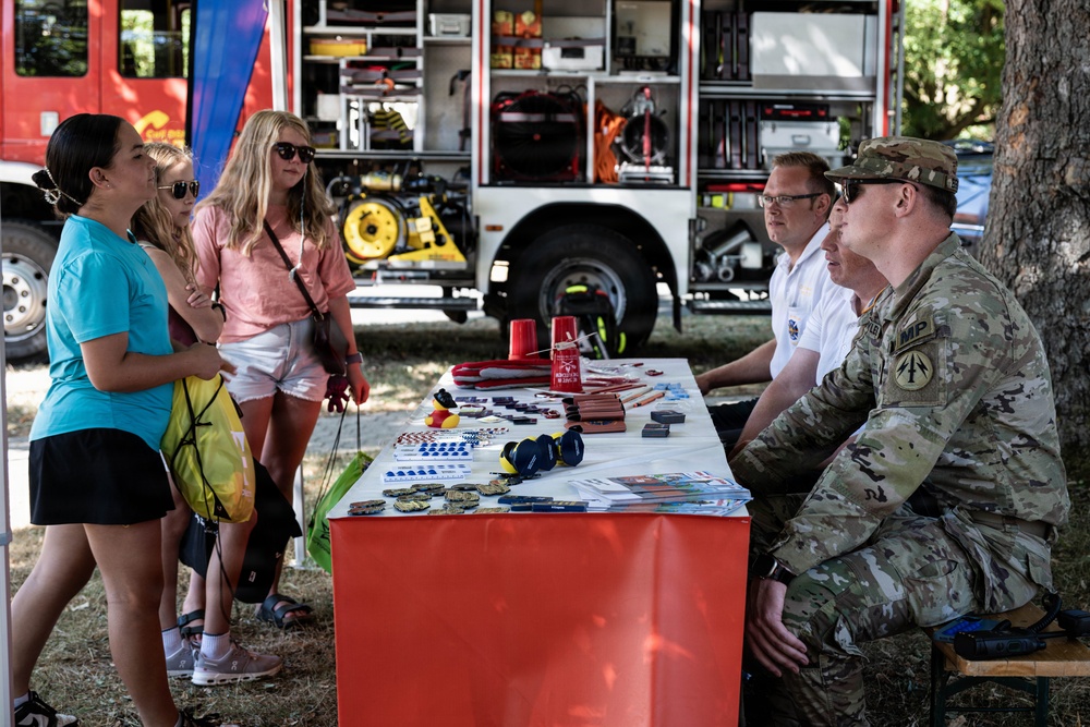 An outdoor community outreach or recruitment event featuring military personnel and civilian staff seated behind a display table with promotional items. Three young women are engaging with the representatives across the table. A fire truck with open compartments is visible in the background, suggesting a public safety or community awareness event.