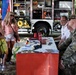 An outdoor community outreach or recruitment event featuring military personnel and civilian staff seated behind a display table with promotional items. Three young women are engaging with the representatives across the table. A fire truck with open compartments is visible in the background, suggesting a public safety or community awareness event.