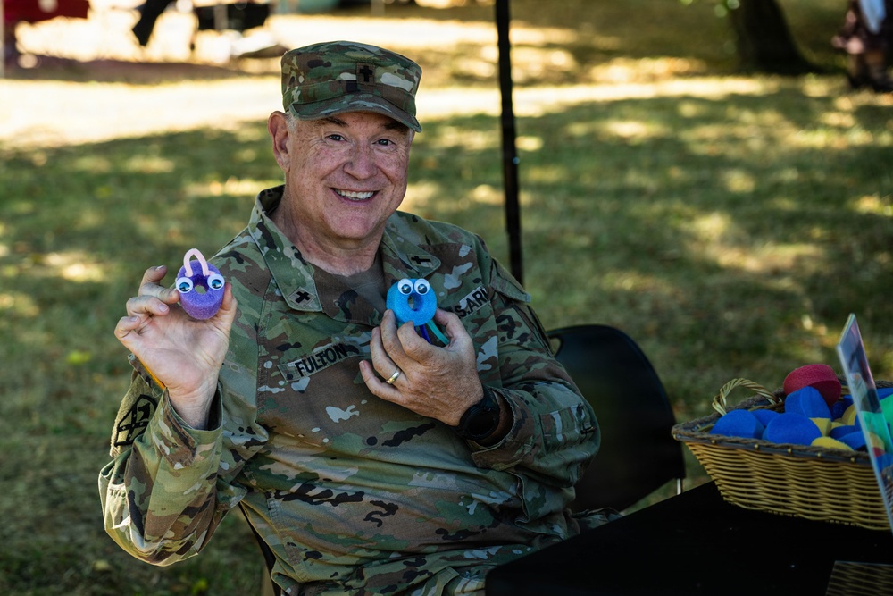 A smiling U.S. Army soldier wearing OCP (Operational Camouflage Pattern) uniform with the name tape 'FULTON' is seated outdoors, holding up two small handmade craft figures with googly eyes. The soldier appears to be at a community or family event, with a wicker basket of colorful foam craft items visible nearby. The atmosphere is cheerful and informal.