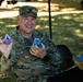 A smiling U.S. Army soldier wearing OCP (Operational Camouflage Pattern) uniform with the name tape 'FULTON' is seated outdoors, holding up two small handmade craft figures with googly eyes. The soldier appears to be at a community or family event, with a wicker basket of colorful foam craft items visible nearby. The atmosphere is cheerful and informal.