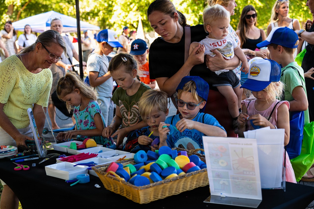 An outdoor community event or fair where children and adults are gathered around a craft activity table. A woman in black holds a toddler while supervising several young children engaged in a hands-on craft project. The setting appears to be a park or outdoor venue with tents and trees visible in the background.