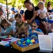 An outdoor community event or fair where children and adults are gathered around a craft activity table. A woman in black holds a toddler while supervising several young children engaged in a hands-on craft project. The setting appears to be a park or outdoor venue with tents and trees visible in the background.