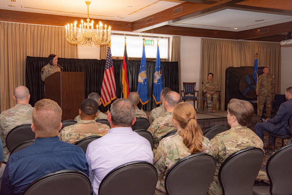 A military ceremony or briefing is taking place in a formal indoor venue, likely a conference or banquet room on a military installation. A female service member in camouflage uniform is speaking at a podium while an audience of predominantly uniformed military personnel listens. Flags including the United States flag, German flag, and U.S. Air Force flags are prominently displayed on the stage.