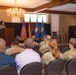A military ceremony or briefing is taking place in a formal indoor venue, likely a conference or banquet room on a military installation. A female service member in camouflage uniform is speaking at a podium while an audience of predominantly uniformed military personnel listens. Flags including the United States flag, German flag, and U.S. Air Force flags are prominently displayed on the stage.