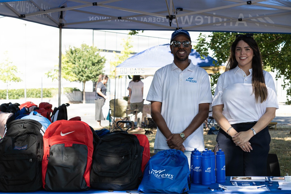 Two representatives from Andrews Federal Credit Union are staffing an outdoor promotional booth at what appears to be a community or campus event. The table displays branded giveaway items including blue drawstring bags, water bottles, and brochures, alongside a collection of backpacks. Both staff members are wearing white Andrews Federal Credit Union polo shirts and smiling at the camera.