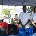 Two representatives from Andrews Federal Credit Union are staffing an outdoor promotional booth at what appears to be a community or campus event. The table displays branded giveaway items including blue drawstring bags, water bottles, and brochures, alongside a collection of backpacks. Both staff members are wearing white Andrews Federal Credit Union polo shirts and smiling at the camera.