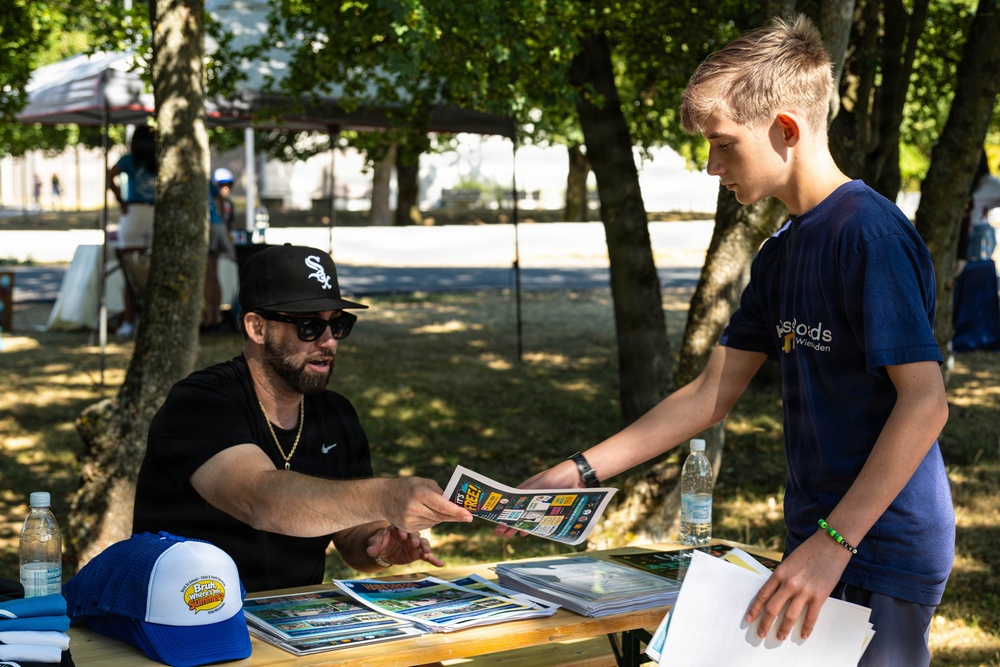 A man wearing a Chicago White Sox cap and sunglasses sits at an outdoor table, handing a booklet or pamphlet to a young teenage boy at what appears to be a community event or fair. The setting is a shaded outdoor area with trees, tents, and other attendees visible in the background. The table is covered with stacked printed materials, hats, and water bottles.