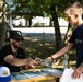 A man wearing a Chicago White Sox cap and sunglasses sits at an outdoor table, handing a booklet or pamphlet to a young teenage boy at what appears to be a community event or fair. The setting is a shaded outdoor area with trees, tents, and other attendees visible in the background. The table is covered with stacked printed materials, hats, and water bottles.