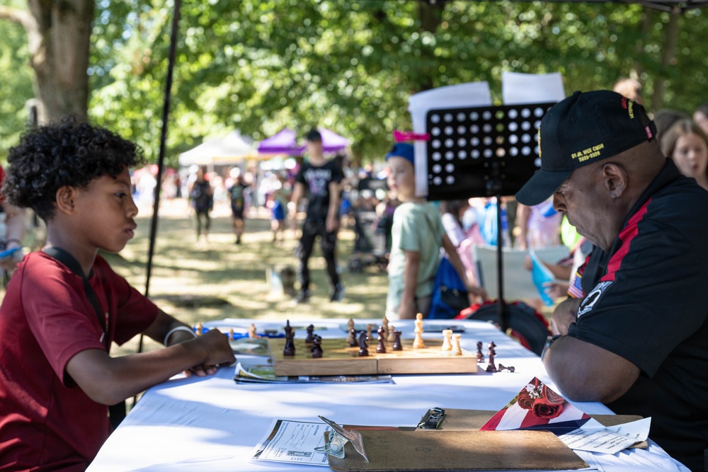 A young boy and an older man are engaged in a chess game at an outdoor community event or festival. They are seated across from each other at a white-clothed table with a wooden chess board between them. The setting appears to be a park with vendor tents and other attendees visible in the background.