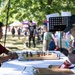 A young boy and an older man are engaged in a chess game at an outdoor community event or festival. They are seated across from each other at a white-clothed table with a wooden chess board between them. The setting appears to be a park with vendor tents and other attendees visible in the background.