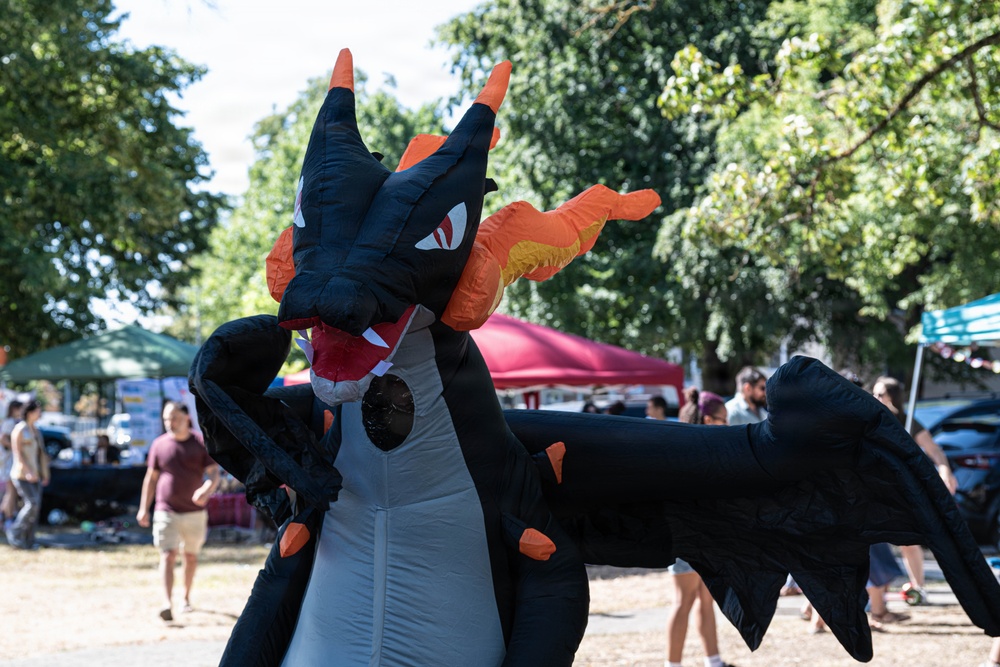 A person wearing a large inflatable dragon costume dominates the foreground at what appears to be an outdoor festival or market event. The costume is black and orange, resembling a fantasy dragon character with wings, horns, and a red mouth. The background shows vendor tents, trees, and attendees in a sunny outdoor setting.