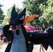 A person wearing a large inflatable dragon costume dominates the foreground at what appears to be an outdoor festival or market event. The costume is black and orange, resembling a fantasy dragon character with wings, horns, and a red mouth. The background shows vendor tents, trees, and attendees in a sunny outdoor setting.