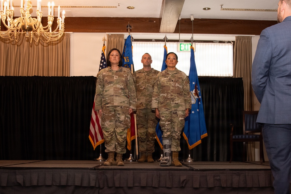 Three military personnel in OCP (Operational Camouflage Pattern) uniforms stand at attention on a raised stage during what appears to be a formal military ceremony. The setting is an indoor banquet or event hall with flags including the United States flag and Air Force flag displayed behind them. A civilian in a blue suit is partially visible on the right edge of the frame.