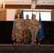 Three military personnel in OCP (Operational Camouflage Pattern) uniforms stand at attention on a raised stage during what appears to be a formal military ceremony. The setting is an indoor banquet or event hall with flags including the United States flag and Air Force flag displayed behind them. A civilian in a blue suit is partially visible on the right edge of the frame.