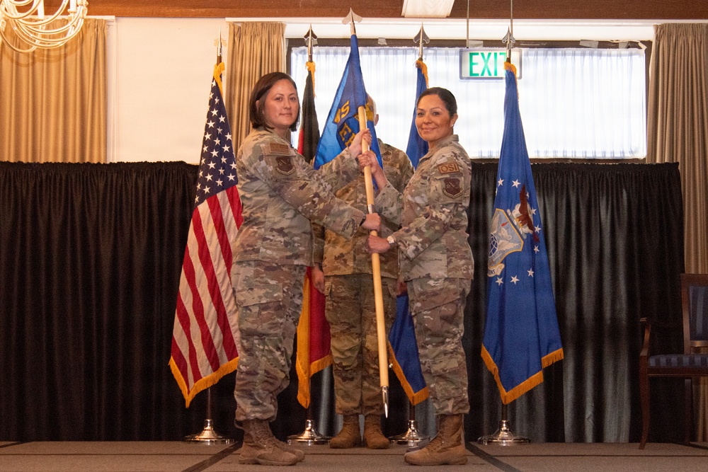 Two female military personnel in U.S. Air Force OCP (Operational Camouflage Pattern) uniforms are participating in a formal change of command ceremony, jointly holding a unit guidon. A third service member stands in the background. The ceremony is taking place in an indoor venue decorated with multiple flags including the U.S. flag, Air Force flag, and what appears to be a German flag.