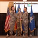Two female military personnel in U.S. Air Force OCP (Operational Camouflage Pattern) uniforms are participating in a formal change of command ceremony, jointly holding a unit guidon. A third service member stands in the background. The ceremony is taking place in an indoor venue decorated with multiple flags including the U.S. flag, Air Force flag, and what appears to be a German flag.