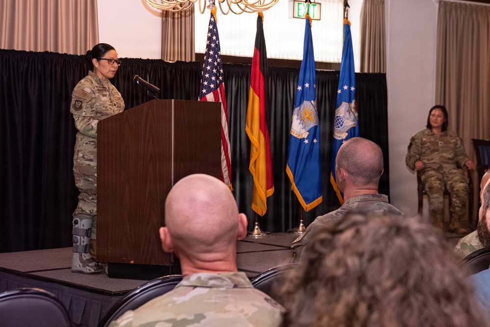 A military ceremony or briefing is taking place in a formal indoor venue. A female service member in camouflage uniform stands at a wooden podium addressing an audience of seated military personnel. Three flags are displayed behind the podium, including the United States flag, the German flag, and a blue military organizational flag.