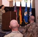 A military ceremony or briefing is taking place in a formal indoor venue. A female service member in camouflage uniform stands at a wooden podium addressing an audience of seated military personnel. Three flags are displayed behind the podium, including the United States flag, the German flag, and a blue military organizational flag.