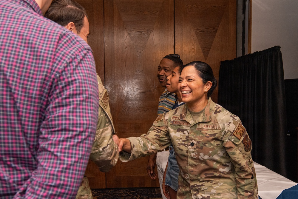 A U.S. Air Force service member in OCP (Operational Camouflage Pattern) uniform is shaking hands with a civilian in a plaid shirt, smiling broadly. The scene appears to take place in an indoor venue with wood-paneled walls, suggesting a formal or ceremonial event. Two additional individuals are visible in the background observing the interaction.