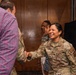 A U.S. Air Force service member in OCP (Operational Camouflage Pattern) uniform is shaking hands with a civilian in a plaid shirt, smiling broadly. The scene appears to take place in an indoor venue with wood-paneled walls, suggesting a formal or ceremonial event. Two additional individuals are visible in the background observing the interaction.